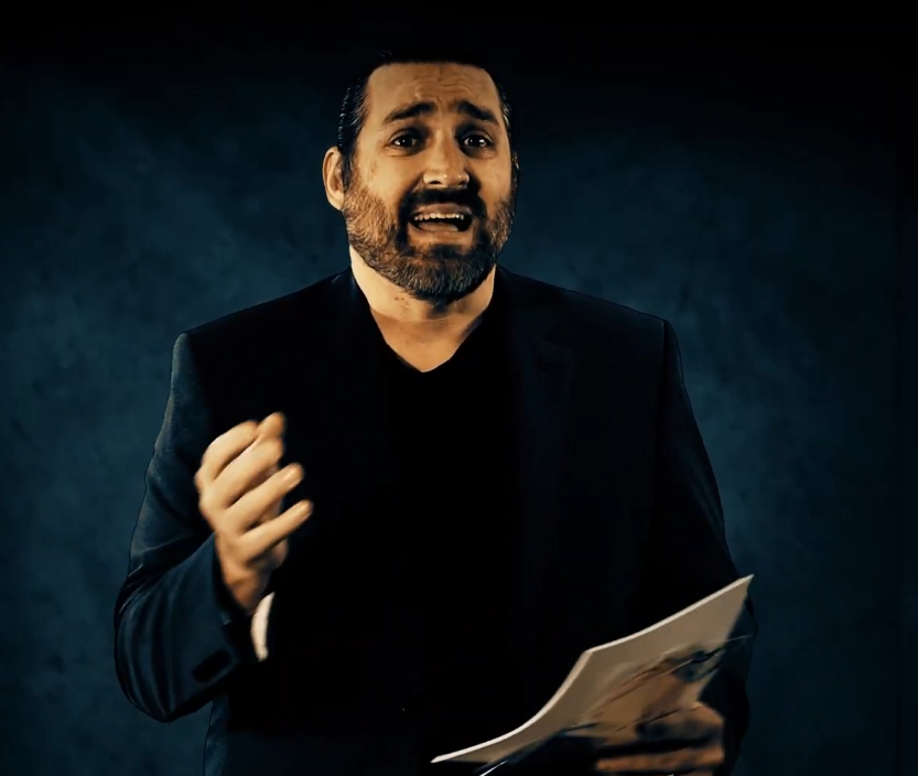 A man with a beard wearing a black suit jacket and black shirt, holding papers and gesturing with his right hand against a dark textured background.