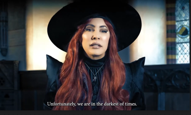 A woman with long red hair wearing a black wide-brim hat and black clothing, shown in a dim interior setting with English subtitles at the bottom reading, 'Unfortunately, we are in the darkest of times.'.