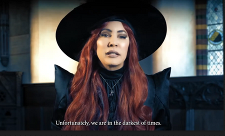 A woman with long red hair wearing a black wide-brim hat and black clothing, shown in a dim interior setting with English subtitles at the bottom reading, 'Unfortunately, we are in the darkest of times.'.