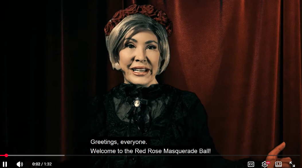 A person in a vintage masquerade costume with a short gray wig, red hat, and black dress stands in front of a red curtain backdrop. Subtitles at the bottom read, 'Greetings, everyone.