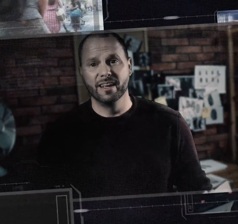 A man with a shaved head and beard wearing a black long-sleeve shirt, standing in front of a brick wall with sketches and a poster pinned on it.