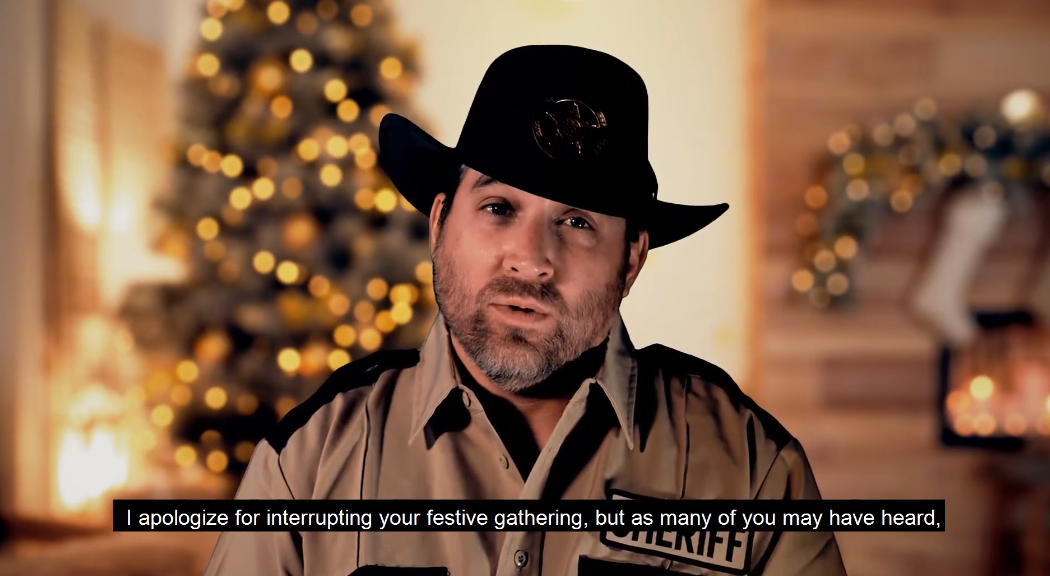 A man wearing a black cowboy hat and a tan sheriff's deputy uniform jacket with 'SHERIFF' on the chest, speaking in front of a warm, blurred holiday background with a Christmas tree and lights.