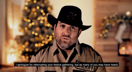 A man wearing a black cowboy hat and a tan sheriff's deputy uniform jacket with 'SHERIFF' on the chest, speaking in front of a warm, blurred holiday background with a Christmas tree and lights.